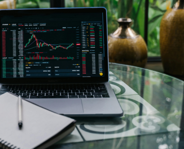 Laptop displaying cryptocurrency trading charts with a notebook and pen on a glass table