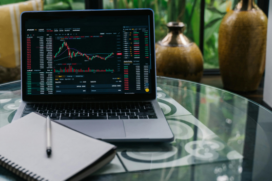 Laptop displaying cryptocurrency trading charts with a notebook and pen on a glass table